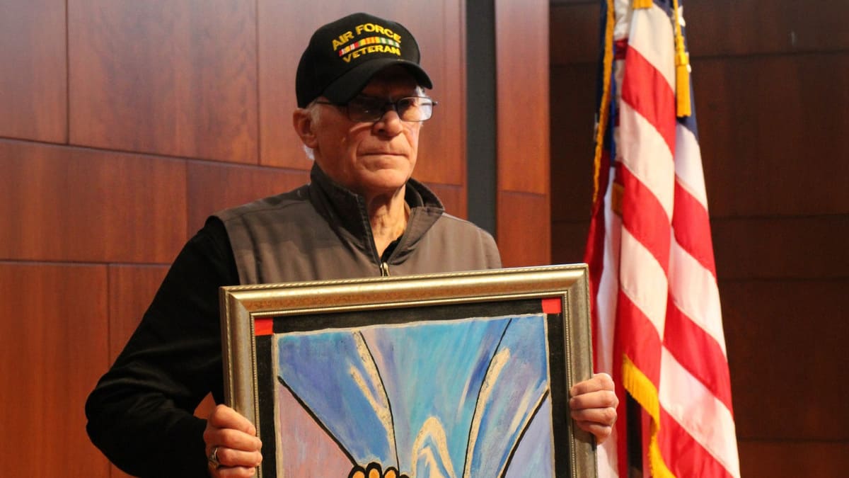 Man wearing an Air Force Veteran hat holding up a painting with US flag in the background.